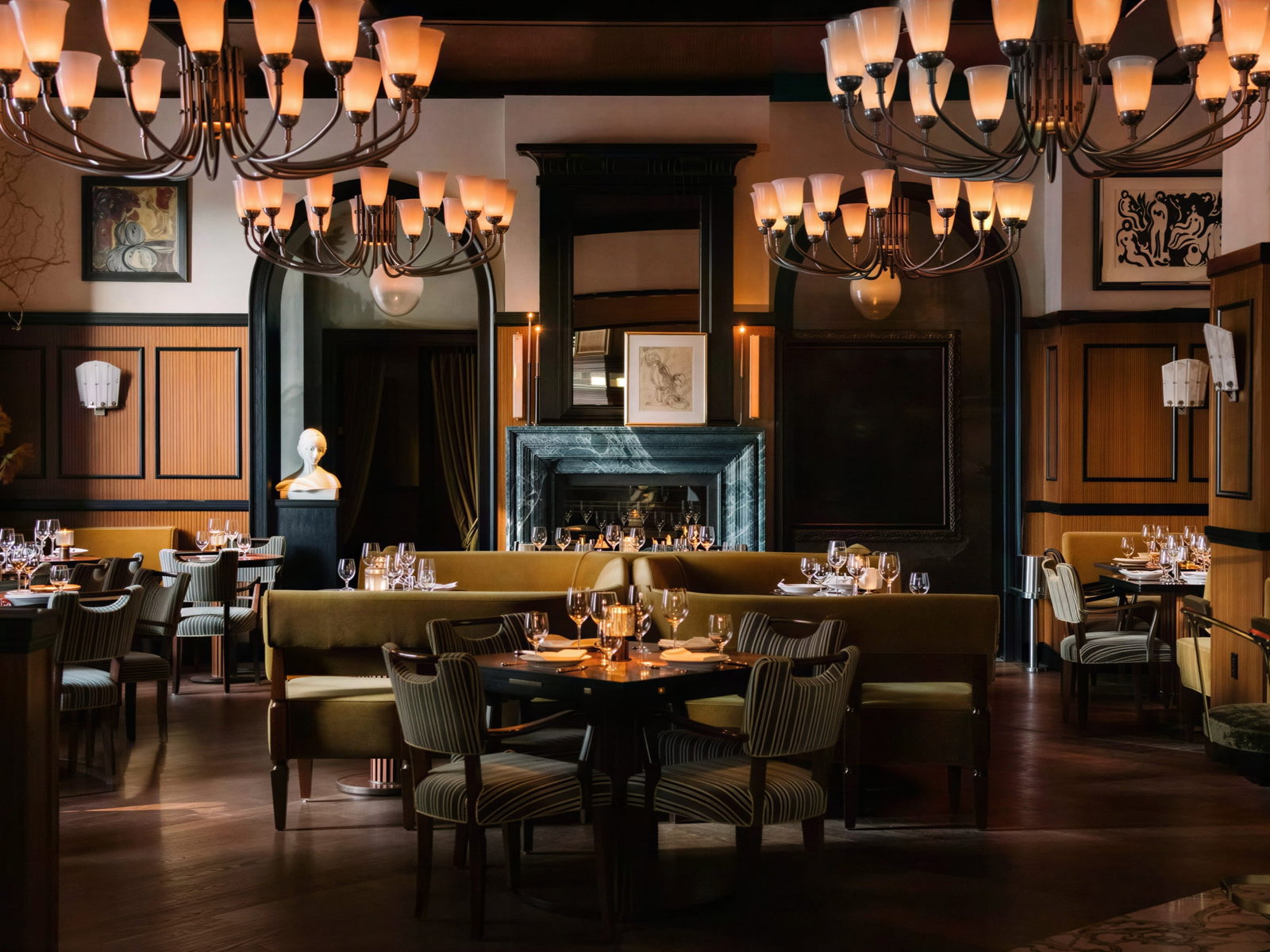 Elegant interior dining room at Locanda Verde Hudson Yards with dim lighting, plush seats, striped chairs, arranged tables with glasses, and large chandeliers, evoking luxury.