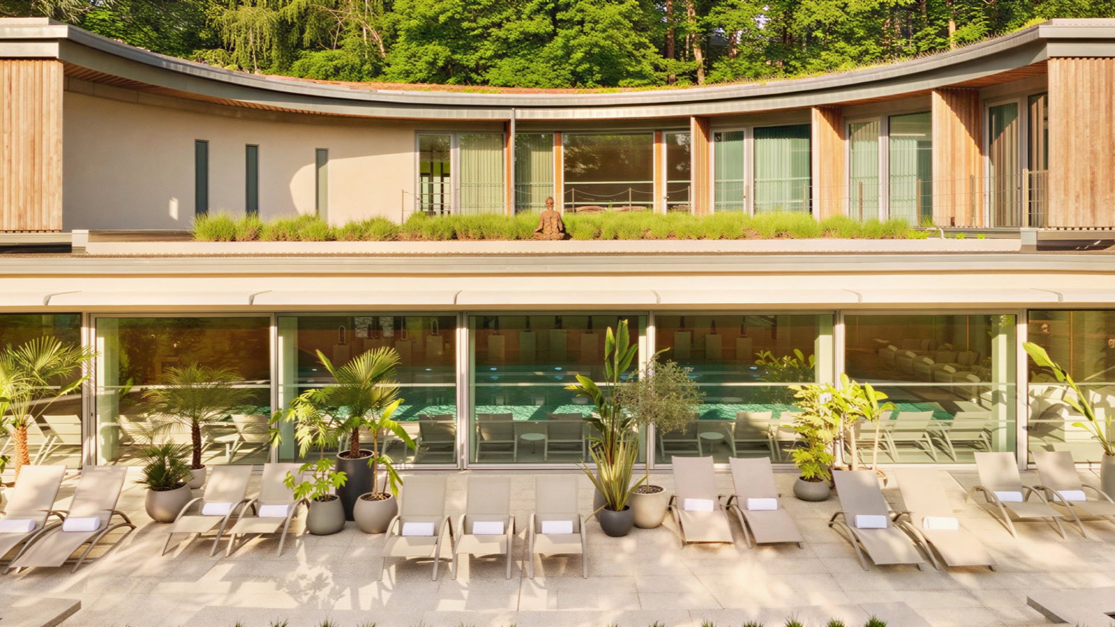 Modern building with a sleek facade, rooftop garden, and poolside lounge chairs. Large windows reflect lush green trees. Serenity prevails.