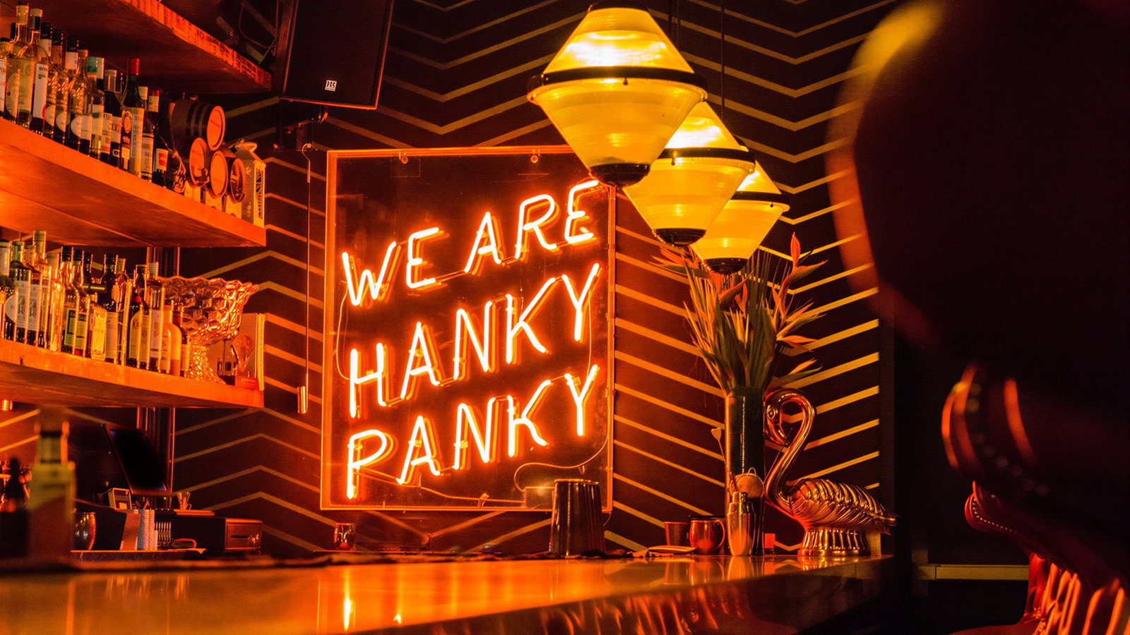 Bar interior with orange neon sign reads "WE ARE HANKY PANKY." Shelves of bottles, striped walls, and warm lighting create a cozy vibe.