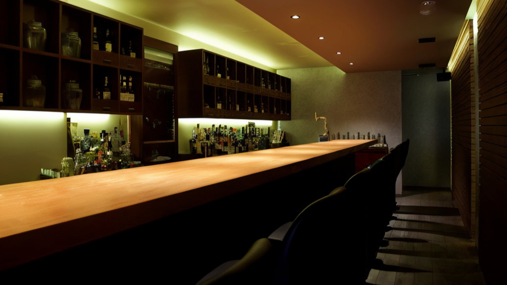 Empty bar with wooden counter, dimly lit shelves of bottles, and black stools. Warm ambiance with sleek, modern design.