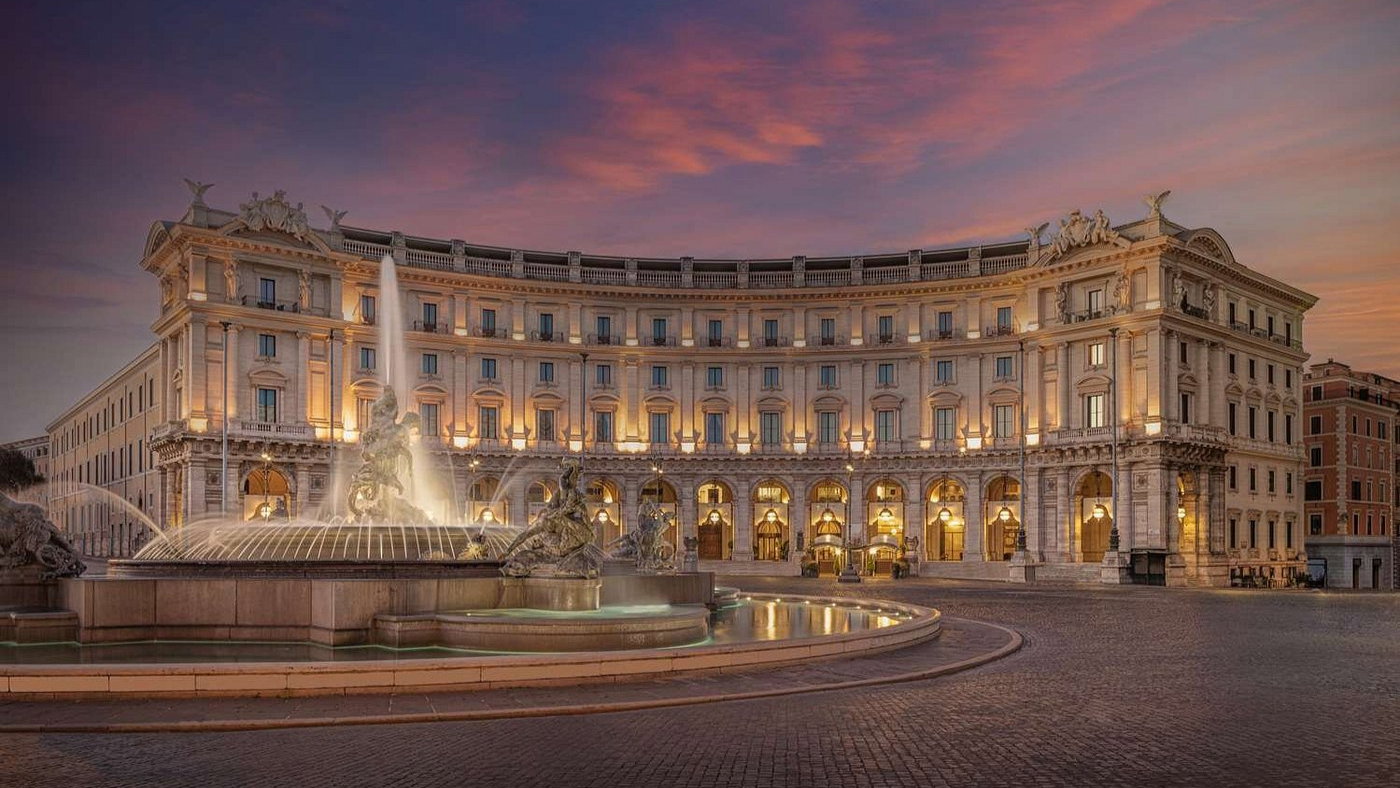 Elegant building facade glows at dusk, with lit arches and a flowing fountain in front. Sky is a gradient of pink and purple hues.