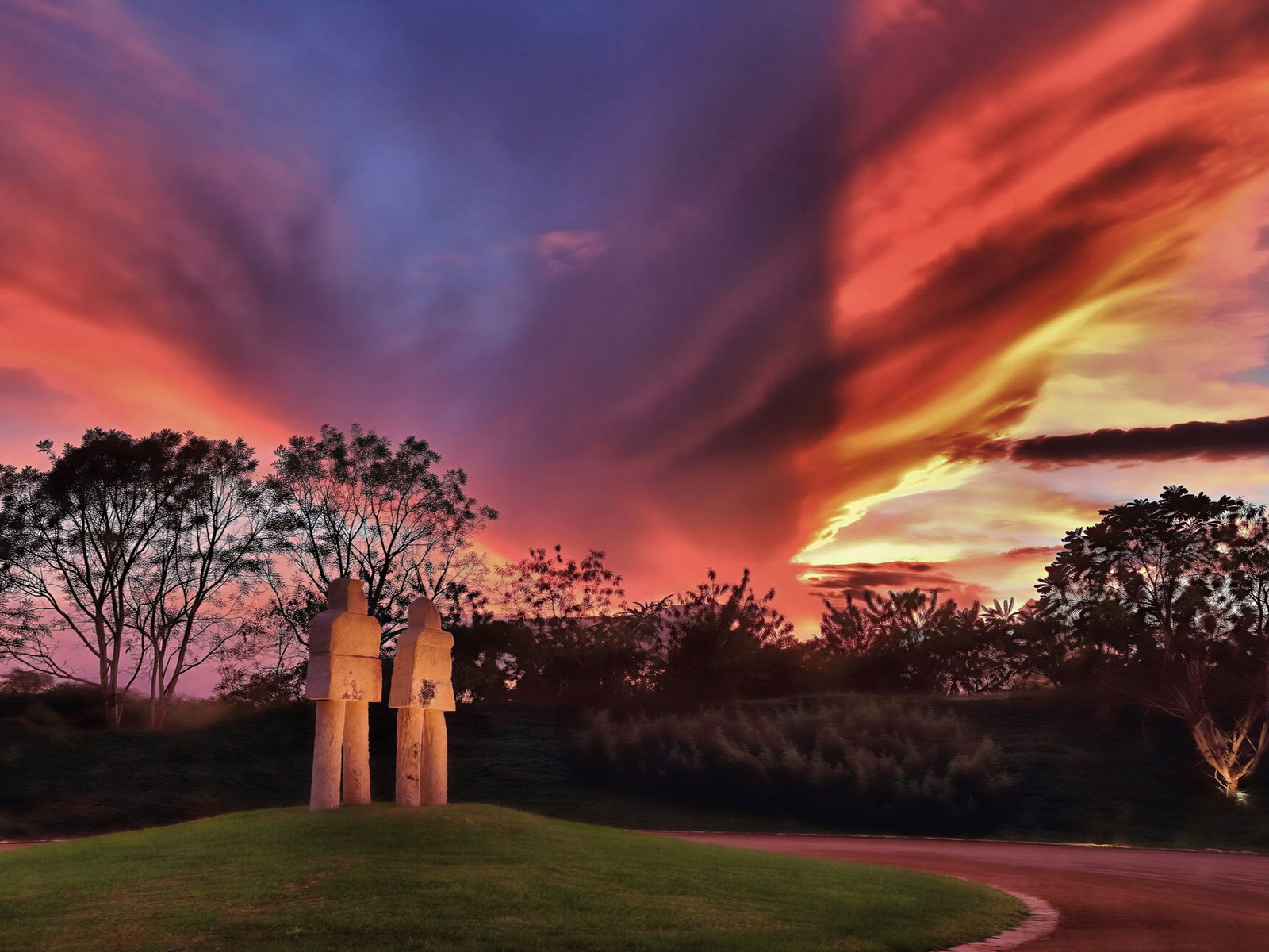 Two stone statues on a grassy hill against a vibrant sunset sky with orange, pink, and purple hues, framed by silhouetted trees.