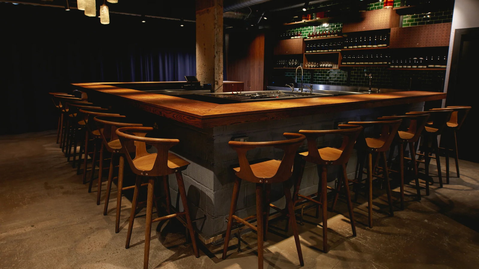 Dimly lit bar with wooden chairs around a U-shaped counter. Green tiled wall and shelves with bottles in the background create a cozy ambiance.