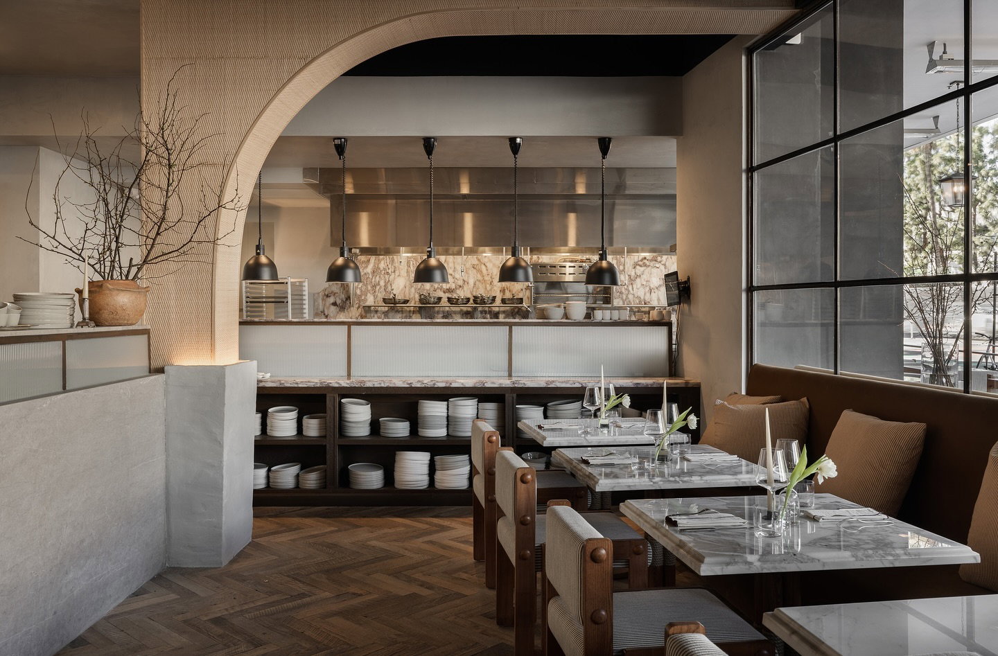 Modern restaurant interior with marble tables, brown chairs, and hanging lamps. Open kitchen view with dishes on shelves. Calm, elegant vibe.