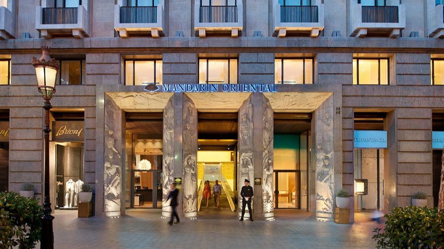 Elegant hotel entrance with stone columns. Sign reads "Mandarin Oriental." Warm lights inside, two people entering, and a guard stands by.