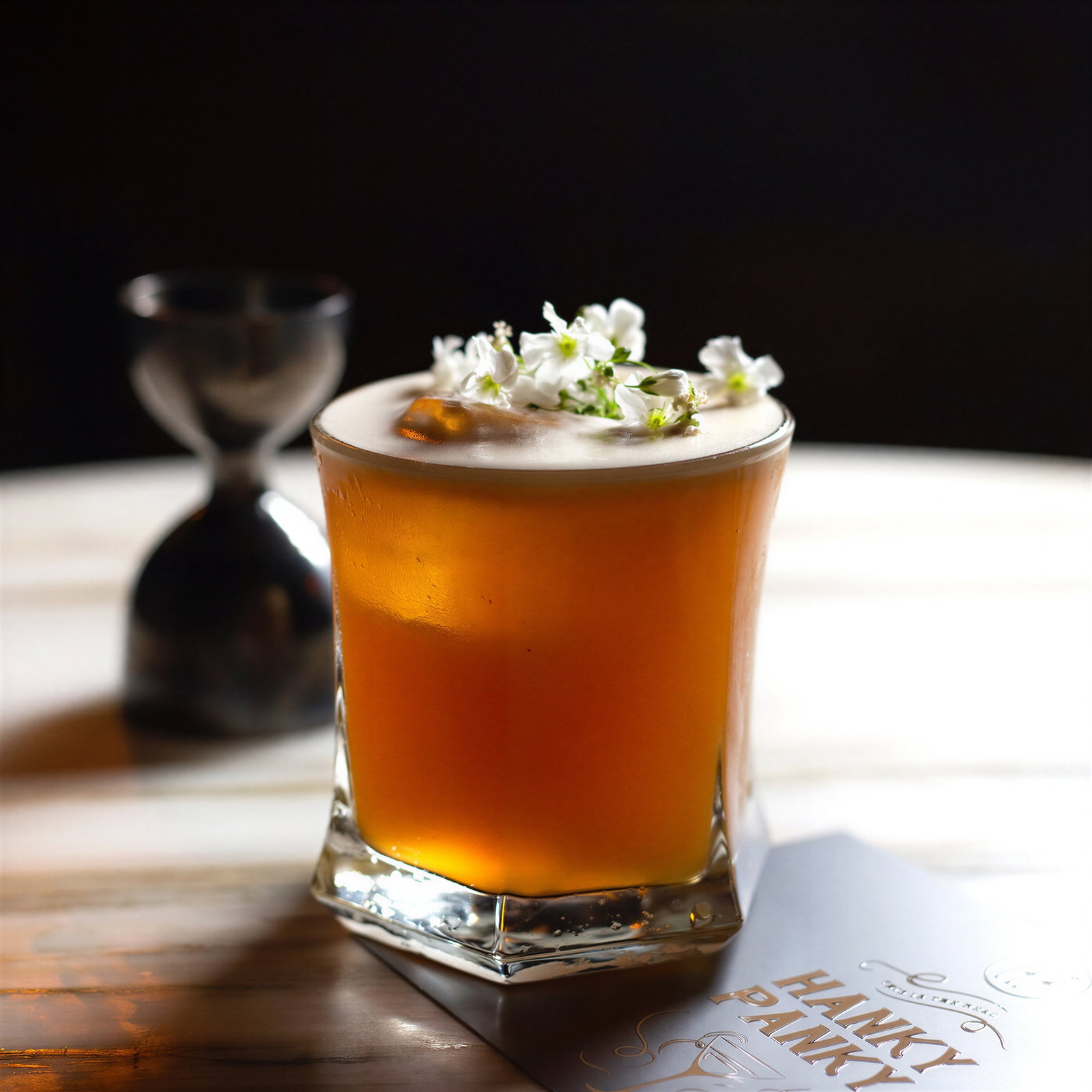 A cocktail garnished with white flowers on a table with a card reading "Hanky Panky;" a metal jigger is blurred in the dark background.