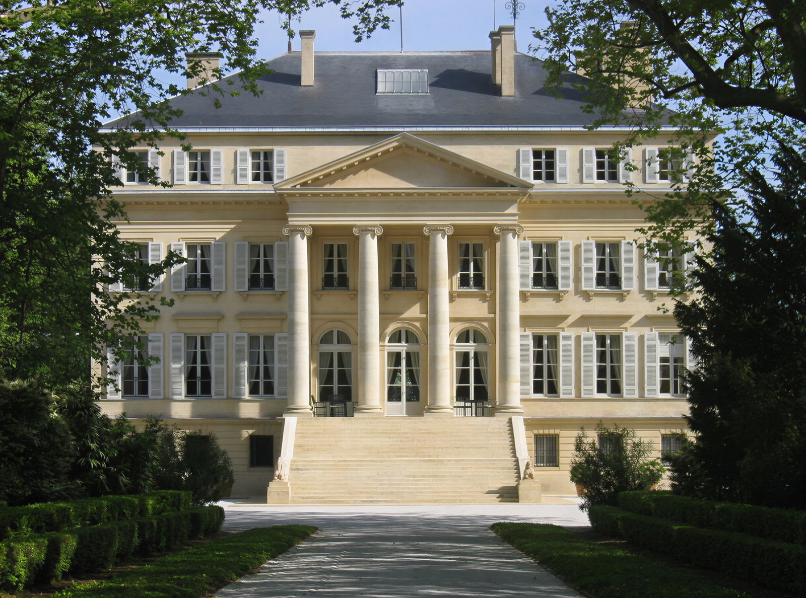 Elegant beige mansion with columns, white shutters, and steps, set in lush green surroundings under a clear blue sky.