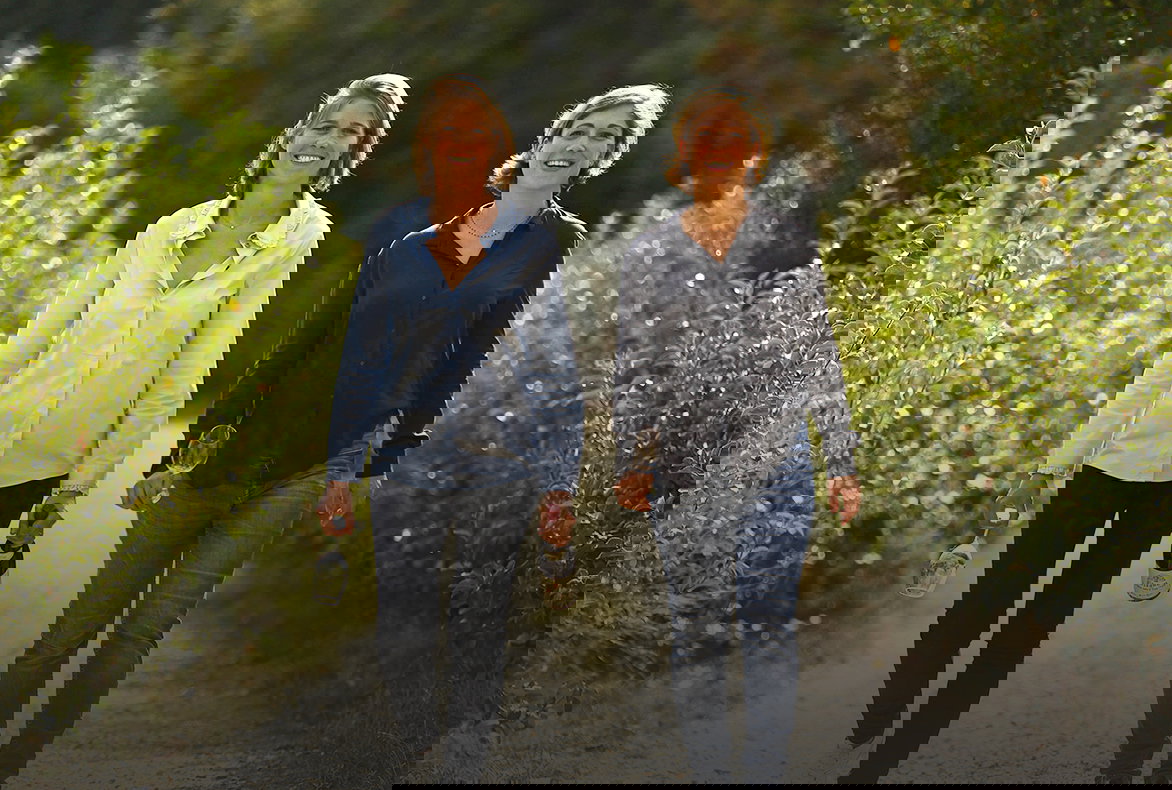 Two women walk on a path, holding wine bottles and glasses, surrounded by lush greenery. They're smiling and wearing casual shirts.