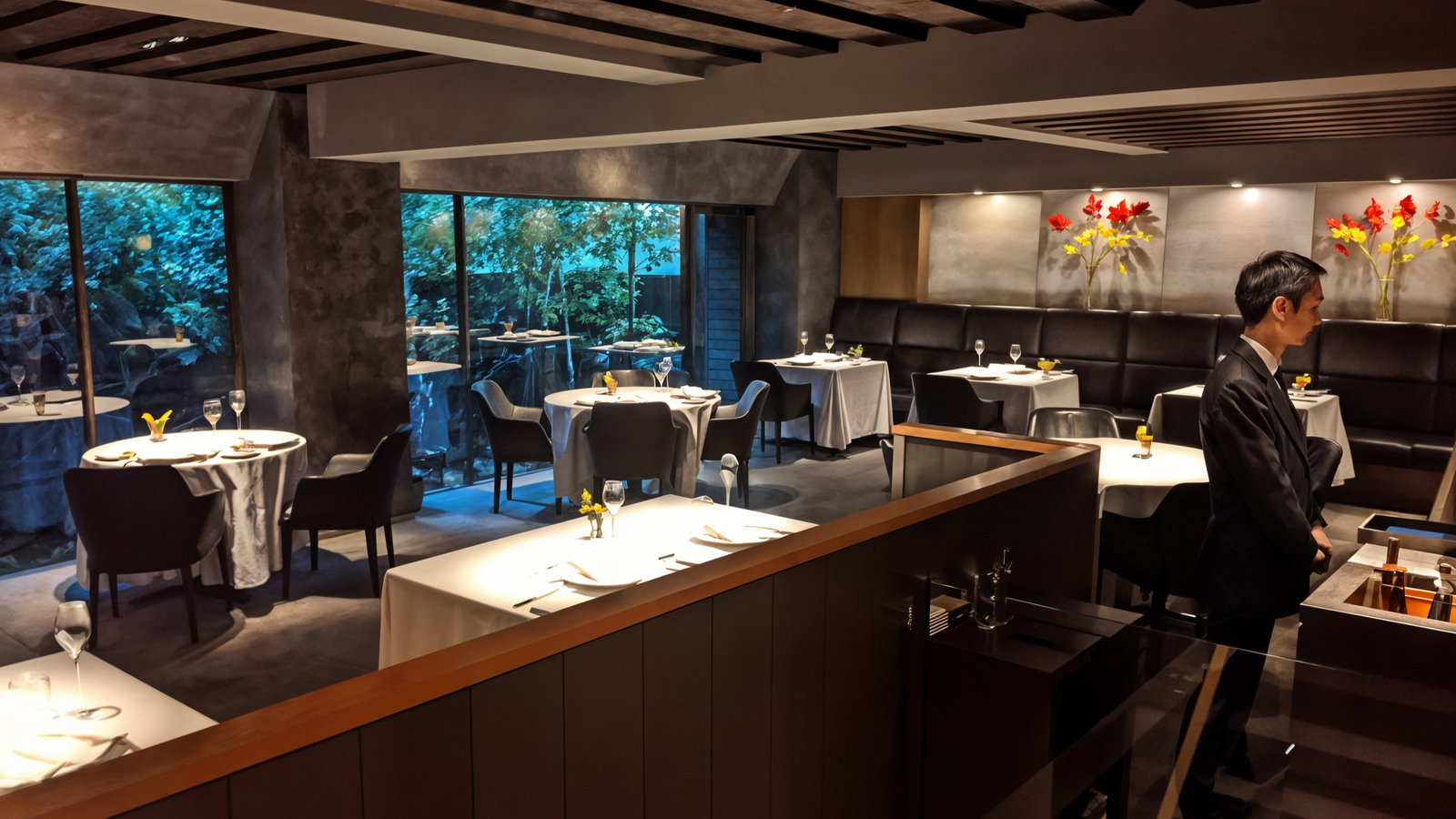 Elegant restaurant interior with neatly set tables, white tablecloths, and floral decor. A waiter stands by. Large windows show greenery outside.