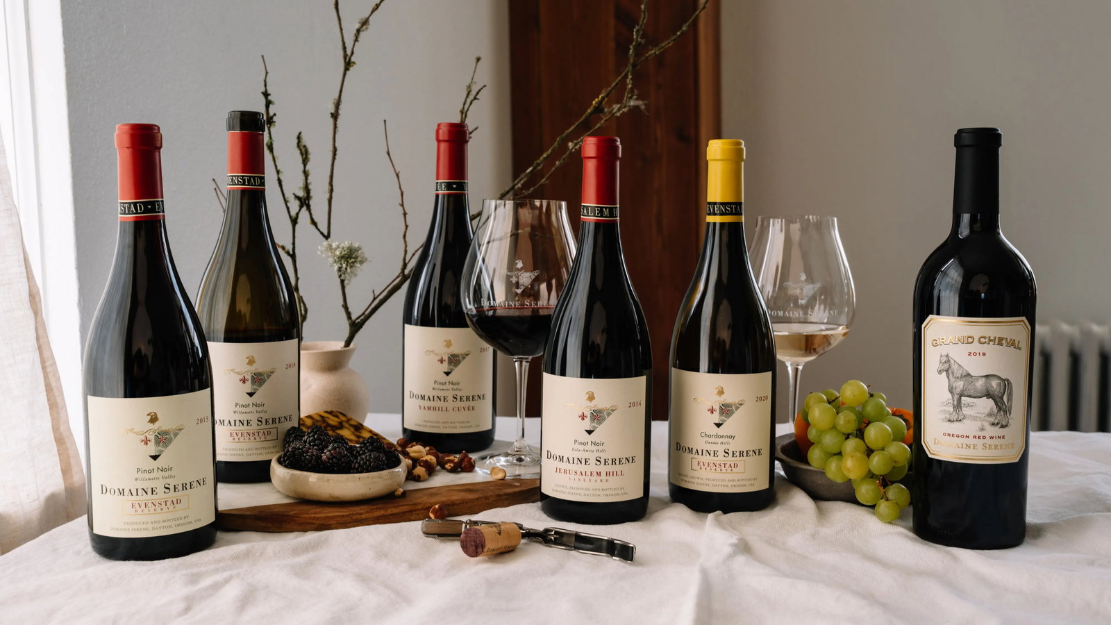 Assorted Domaine Serene wine bottles and glasses on a table with grapes, blackberries, and nuts. Elegant, cozy setting with neutral tones.