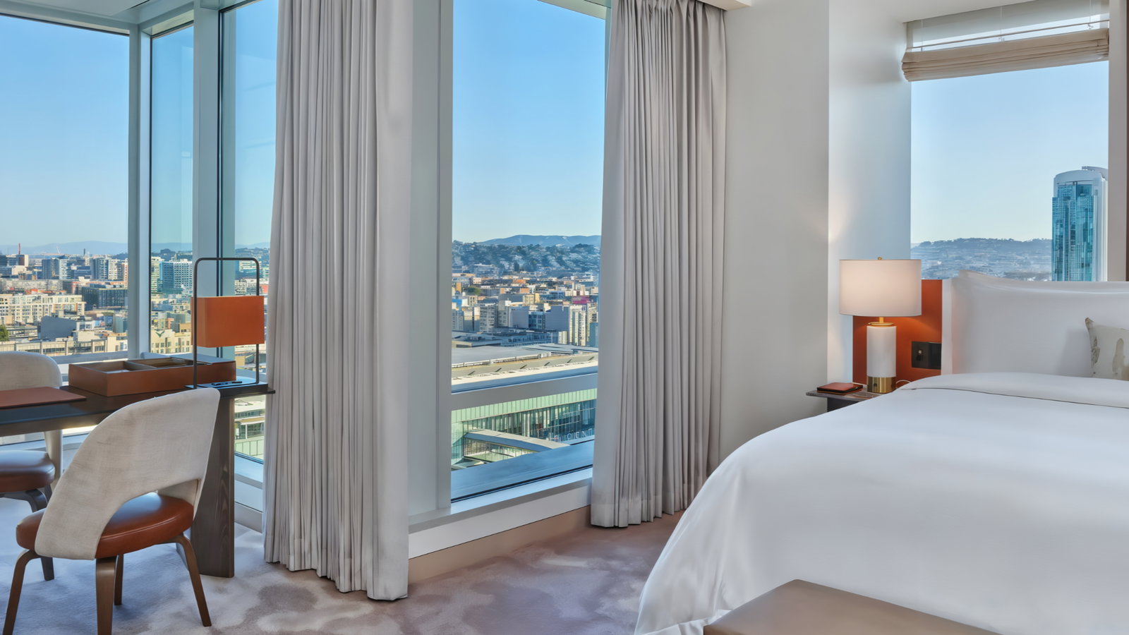 Bright hotel room with a city view from large windows, white bed, orange accents, table with chairs, and soft lighting. Calm, inviting ambiance.