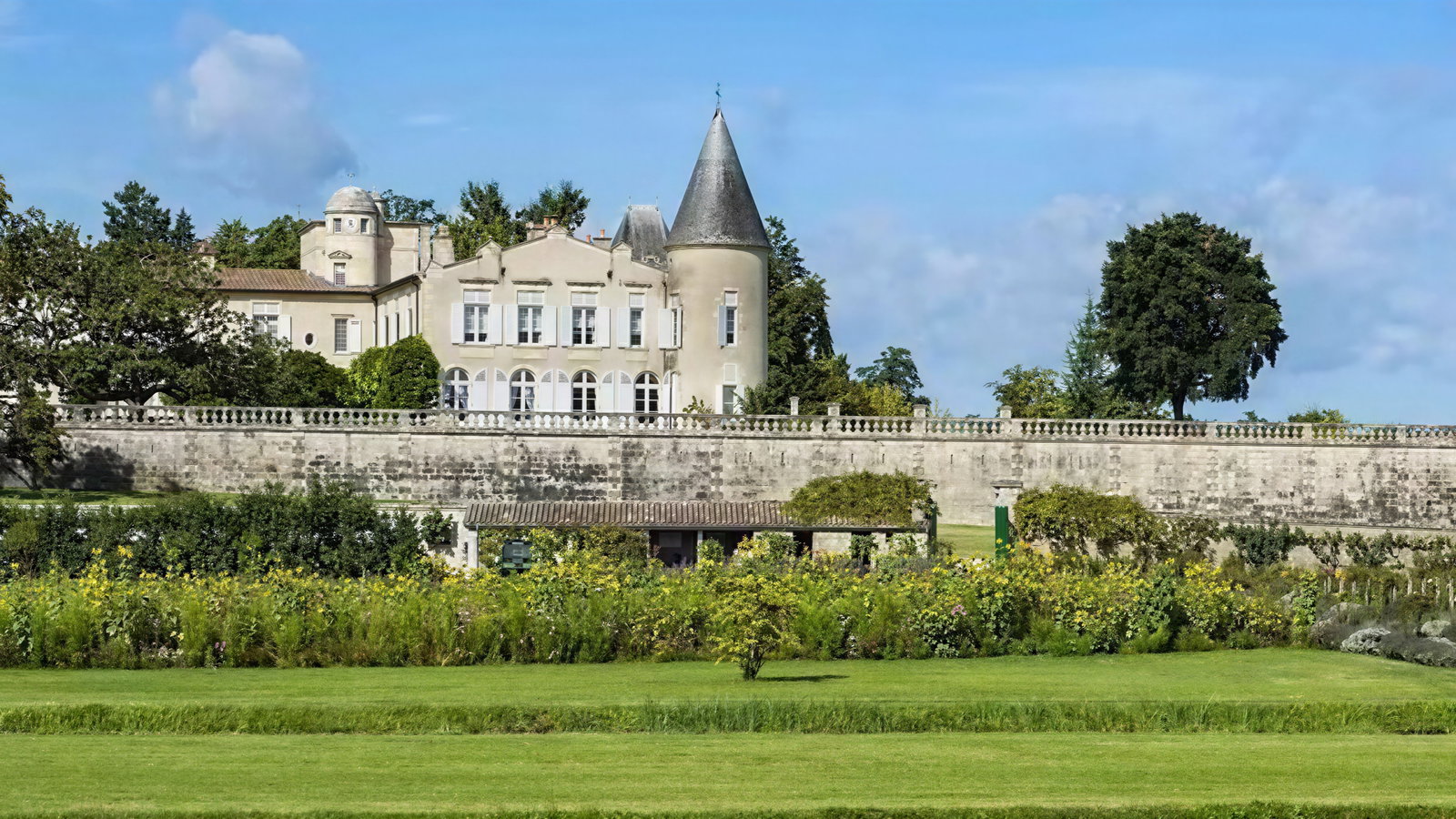 A historic French chateau with conical tower set against a blue sky, surrounded by lush gardens and a stone wall, exuding tranquility.
