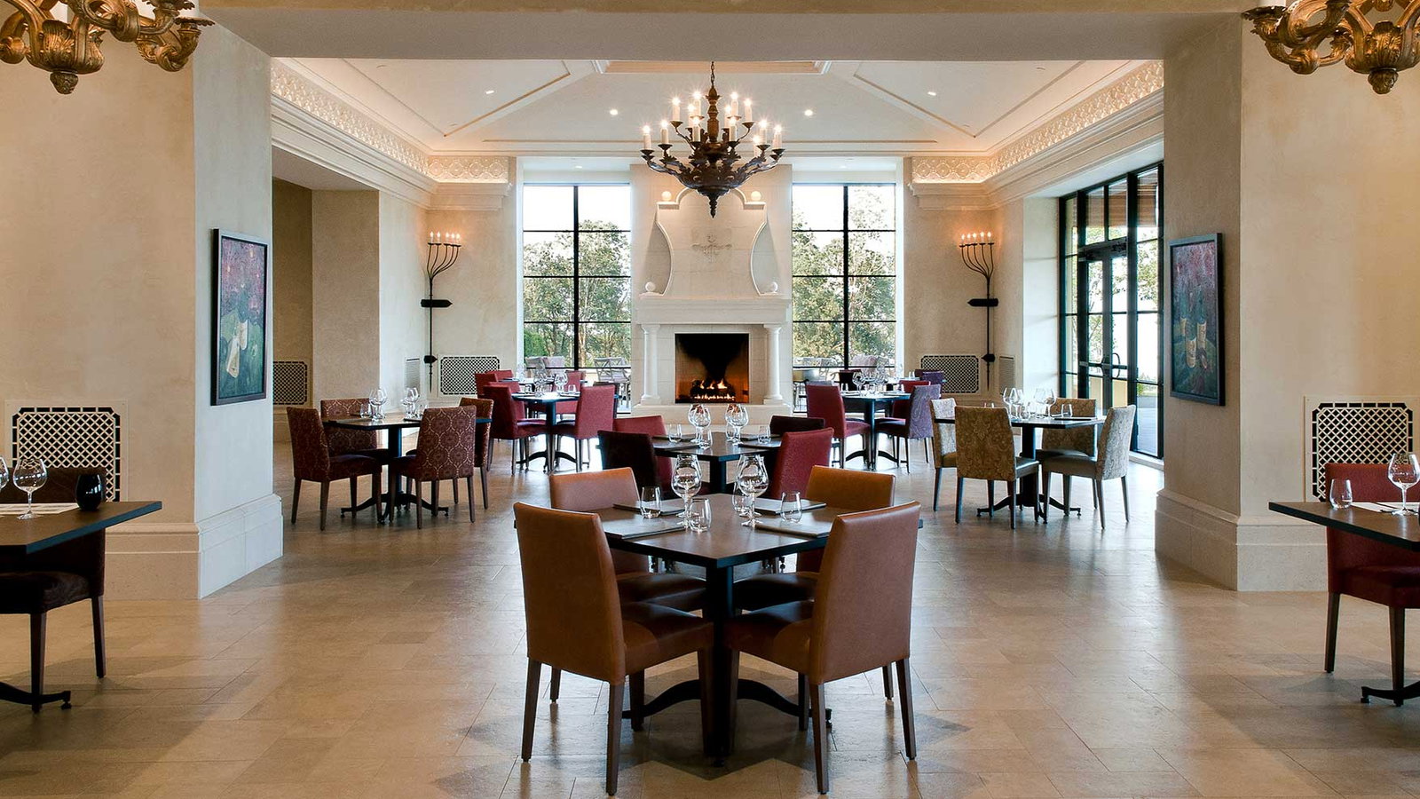 Elegant restaurant interior with empty tables, colorful chairs, and a lit fireplace. Large windows. Warm, inviting ambiance. Chandeliers above.