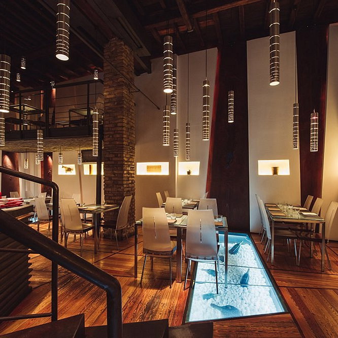 Modern restaurant interior with elegant tables, white chairs, hanging cylindrical lights, wooden floor, and fish visible through a glass floor. Cozy ambiance.