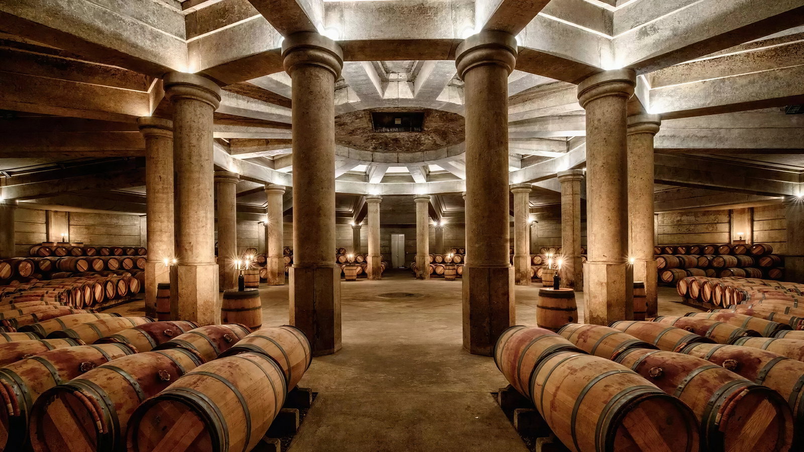Dimly lit wine cellar with rows of wooden barrels and stone columns. Warm lighting creates a cozy, rustic atmosphere. No text visible.