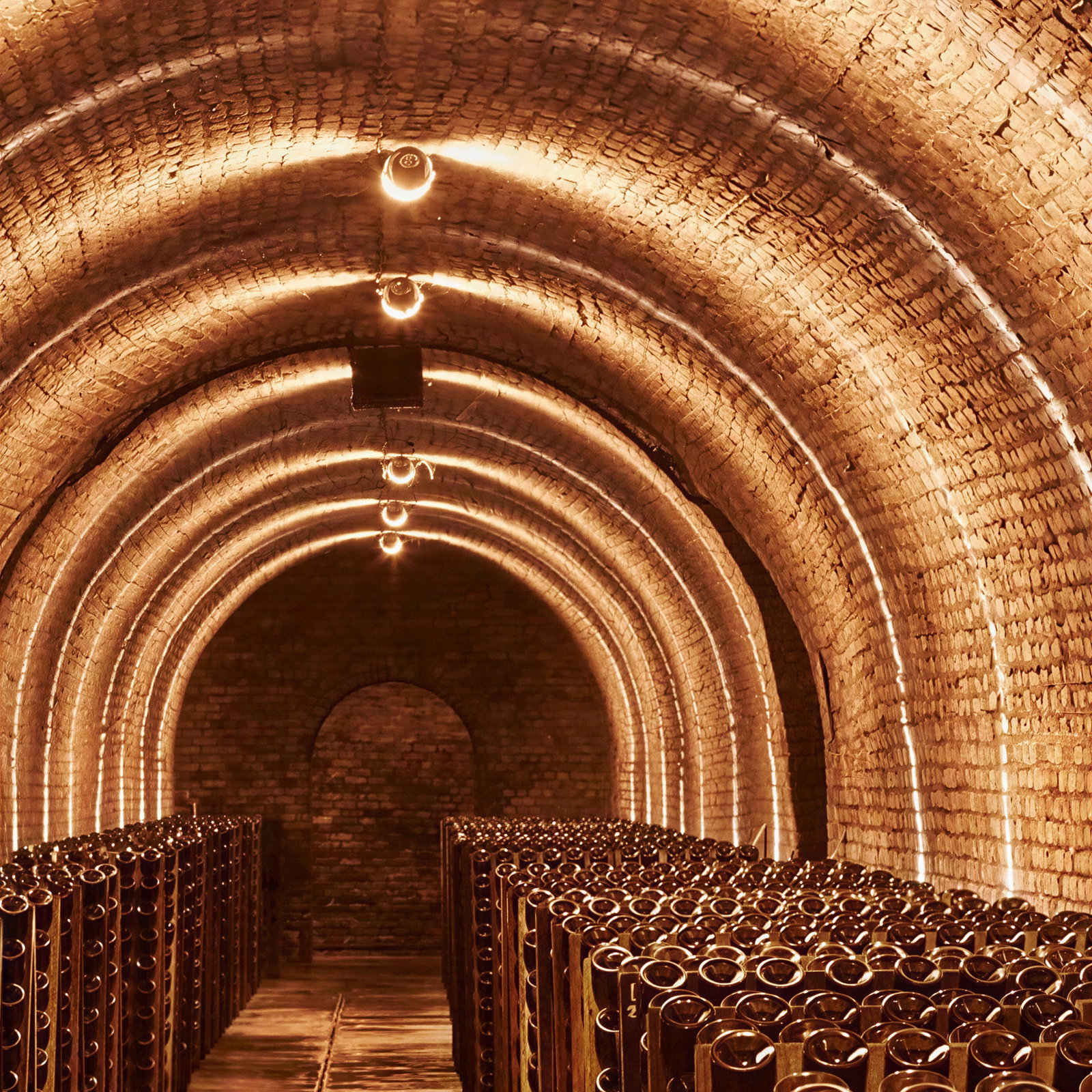 Illuminated brick wine cellar with rows of stacked bottles. Warm lighting creates a cozy atmosphere, highlighting the arched ceiling.
