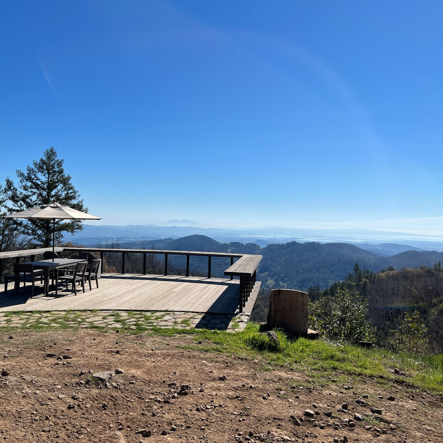 Wooden deck with table, chairs, and an umbrella overlooks a vast hilly landscape under a clear blue sky. Peaceful and scenic setting.