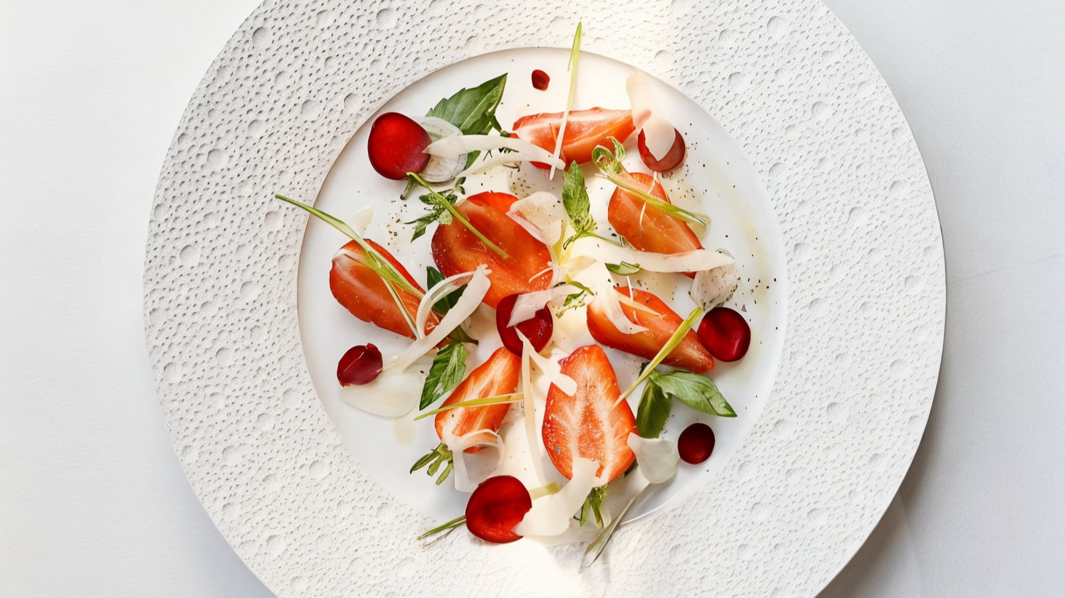 Artfully plated dish with vibrant red tomatoes, herbs, and white ribbons on a textured white plate, creating a fresh and elegant presentation.