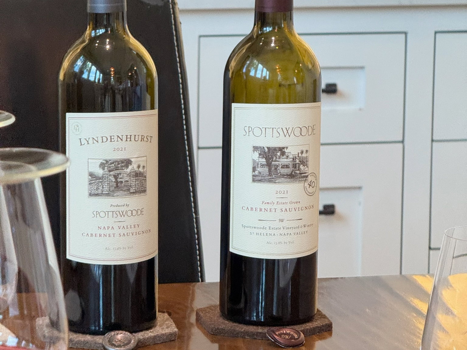Two wine bottles labeled "Lyndenhurst 2021" and "Spottswoode 2021" on a wooden table with glasses, set against a white cabinet backdrop.