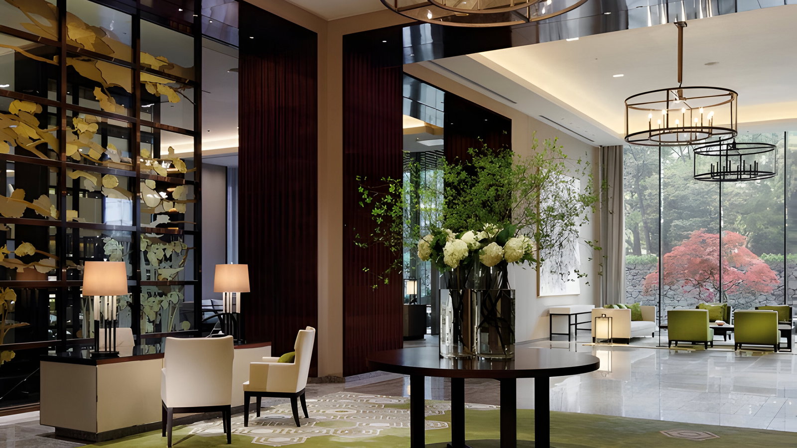 Modern lobby with green chairs, floral decor, and large glass windows showing a garden. Warm lighting and artistic wall accents create a serene atmosphere.