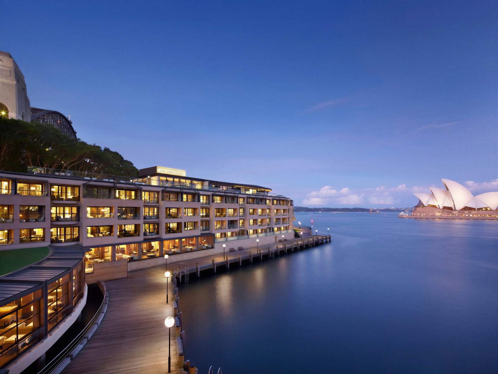 Hotel by the waterfront at dusk, Sydney Opera House in background. Warm lights contrast with blue sky and water, creating a serene mood.