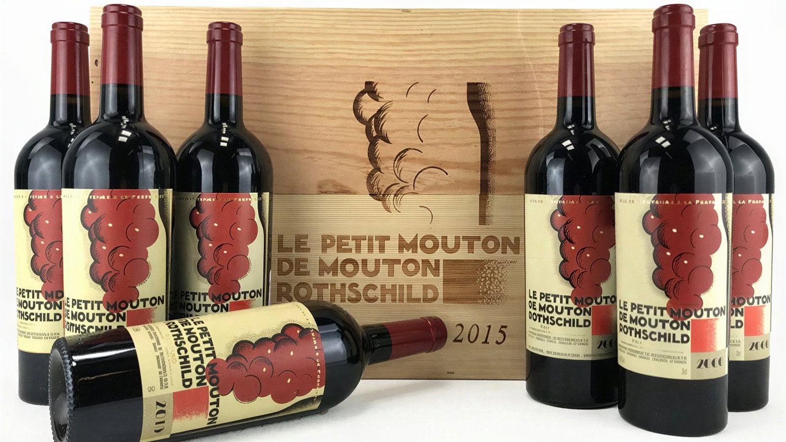 Seven wine bottles with red labels featuring red cloud patterns are arranged before a wooden box labeled "Le Petit Mouton de Mouton Rothschild 2015."