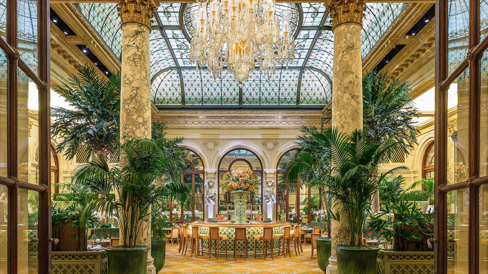 Elegant dining area with large chandeliers, marble columns, lush green plants, and arched windows. Luxurious and serene ambiance.