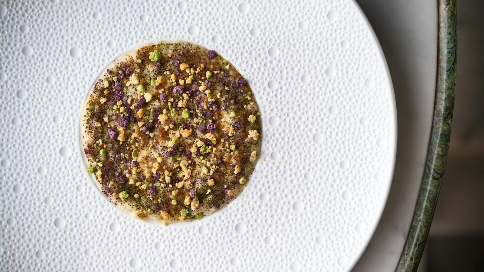 A close-up of a colorful dish with crumbles and herbs on a textured white plate. The background is blurred, creating an elegant presentation.