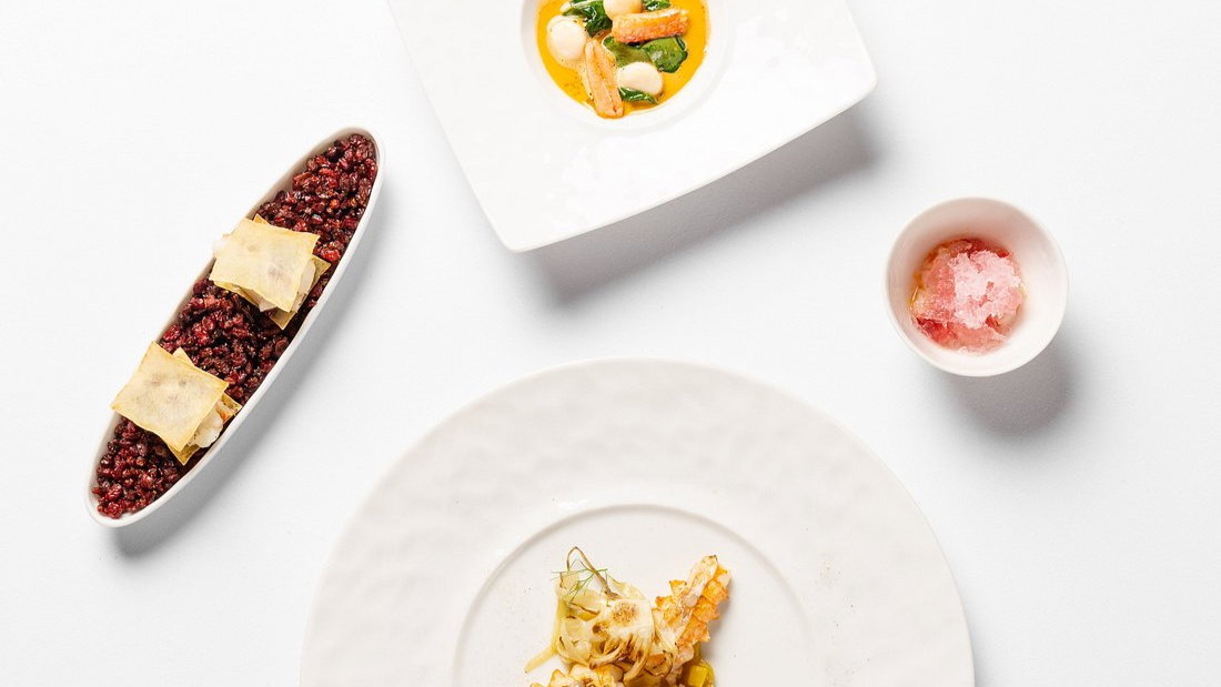Three elegantly presented dishes on a white table. Red grains with crackers, a creamy soup, and a seafood dish. A small cup of pink granita.