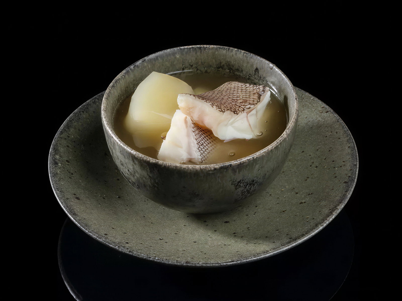 A textured bowl with fish soup, featuring chunks of fish and potatoes in broth. The bowl and plate are gray, set against a black background.