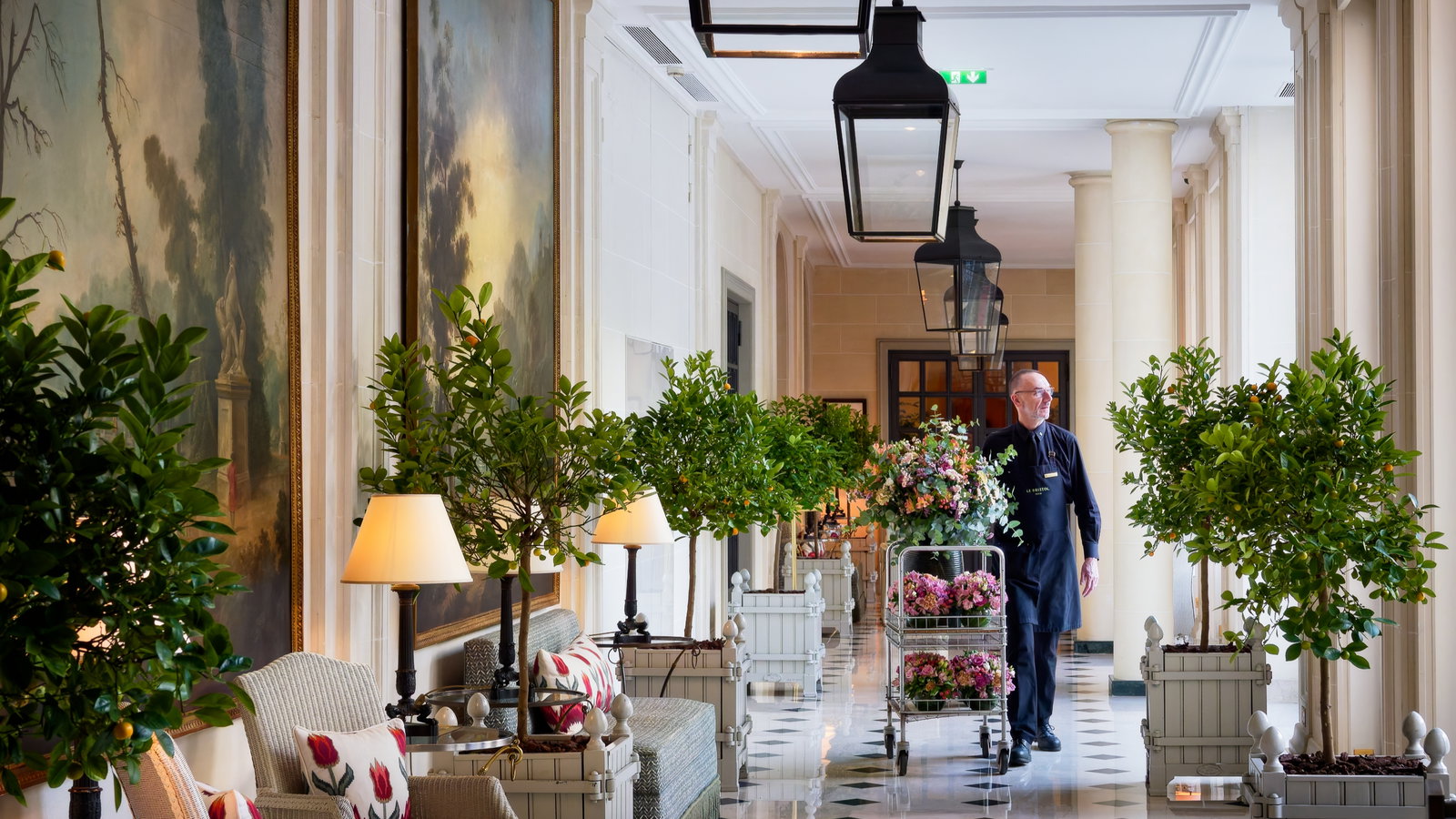 Elegant hallway with a person in uniform pushing a cart of flowers. Lush plants, large paintings, and soft lighting create a serene ambiance.