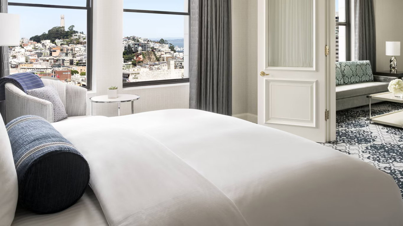 Elegant hotel room with a white bed, blue accents, and a city view through large windows. Open door leads to a stylish lounge area.