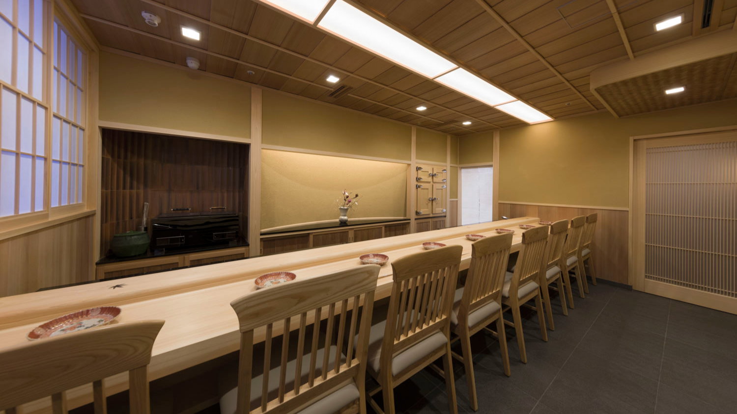 Wooden sushi bar with empty chairs, beige walls, and soft lighting. Plates and utensils set on the counter. Calm, minimalist setting.
