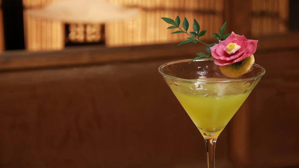 A yellow cocktail in a glass adorned with a pink flower and green leaves, set against a warm wooden background with a soft focus effect.