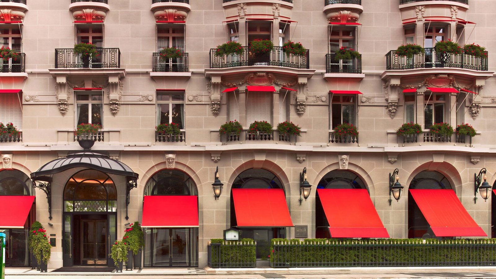 Elegant Hôtel Plaza Athénée exterior facade with red awnings, ornate balconies, and lush greenery. Symmetrical windows enhance the classic architectural design.