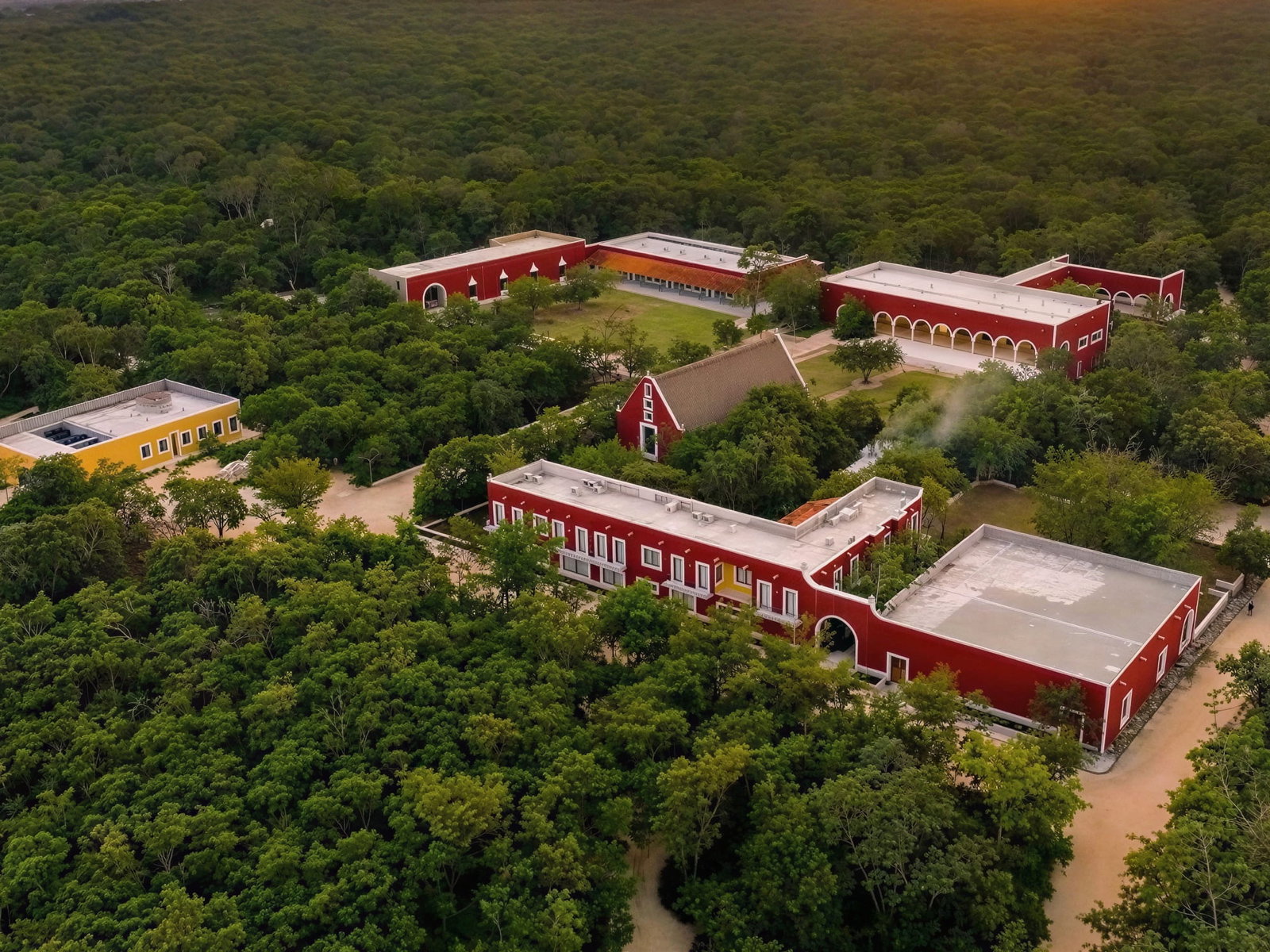 Aerial view of Wakax Hacienda's red and yellow buildings surrounded by lush green forest, under a clear sky. The scene is calm and serene.