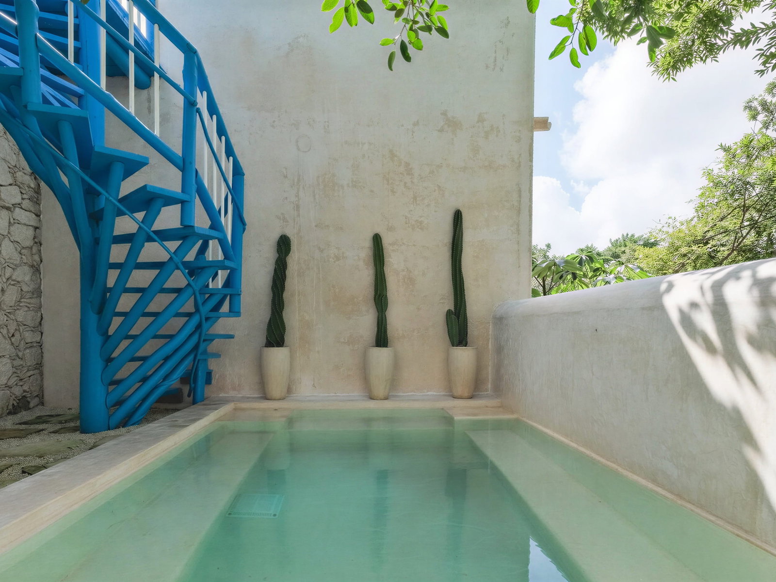 Blue spiral staircase beside the master villa plunge pool at Wakax Hacienda, with three potted cacti against a beige wall. Green trees and a clear sky in the background.
