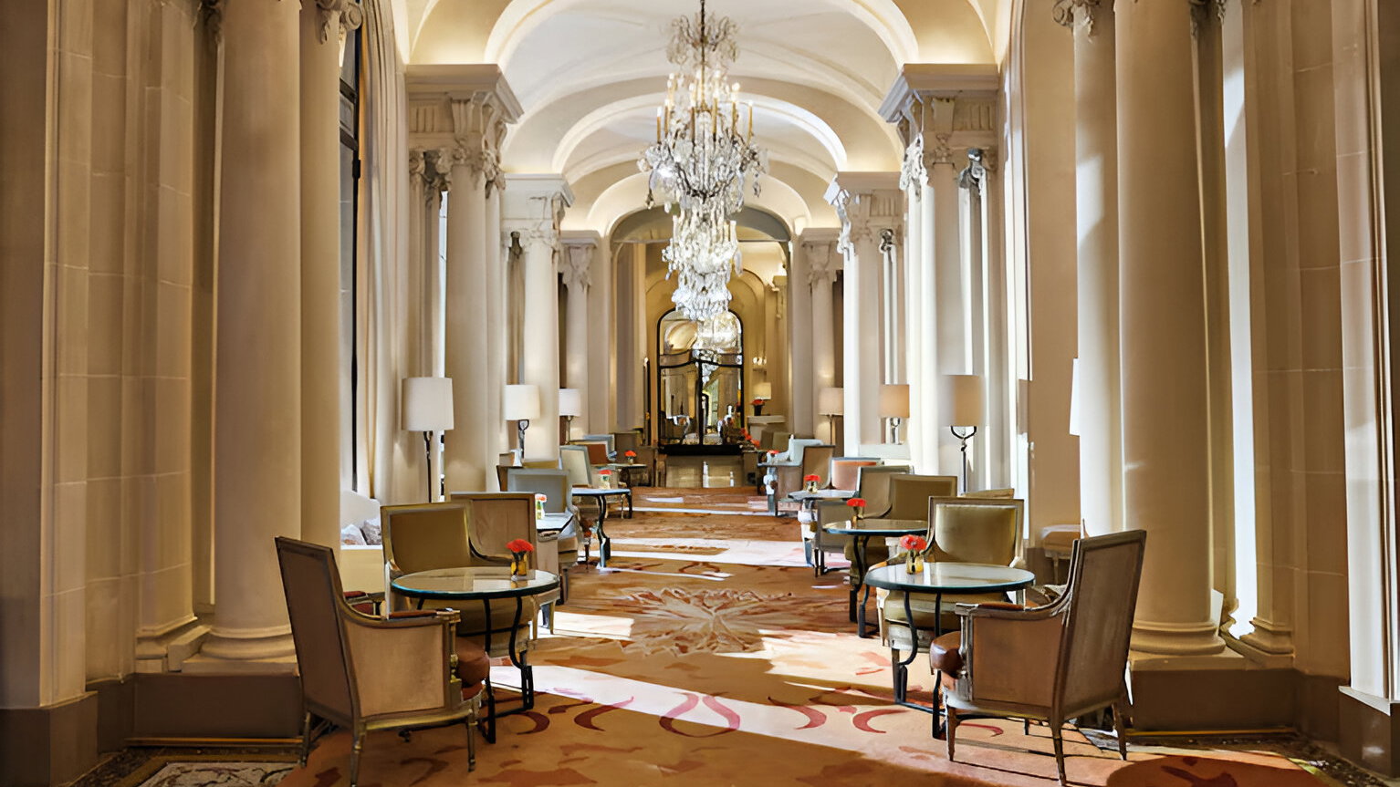 Elegant hotel hallway with chandeliers, high ceilings, and columns. Tables and chairs with red flowers on a patterned carpet enhance the decor.