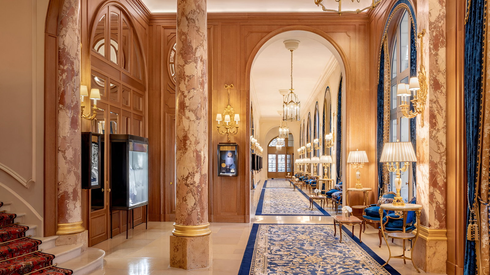 Luxurious hotel lobby with ornate marble pillars, gold accents, and blue velvet chairs. Elegant chandeliers illuminate the patterned carpet.