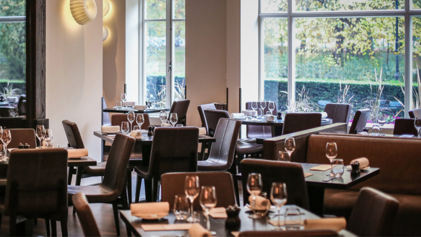 Elegant restaurant with brown leather chairs, neatly set tables with wine glasses, and large windows showing green foliage outside. Peaceful atmosphere.