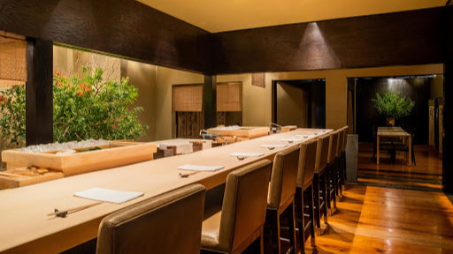 Elegant sushi bar with wooden counters and chairs, warmly lit. Greenery and a serene ambiance create a tranquil dining setting.