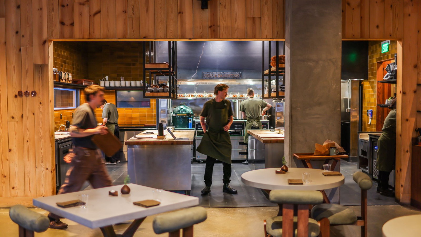 Chefs in dark aprons work in a modern kitchen with wooden walls and orange tiles. One chef moves, creating a dynamic, busy atmosphere.