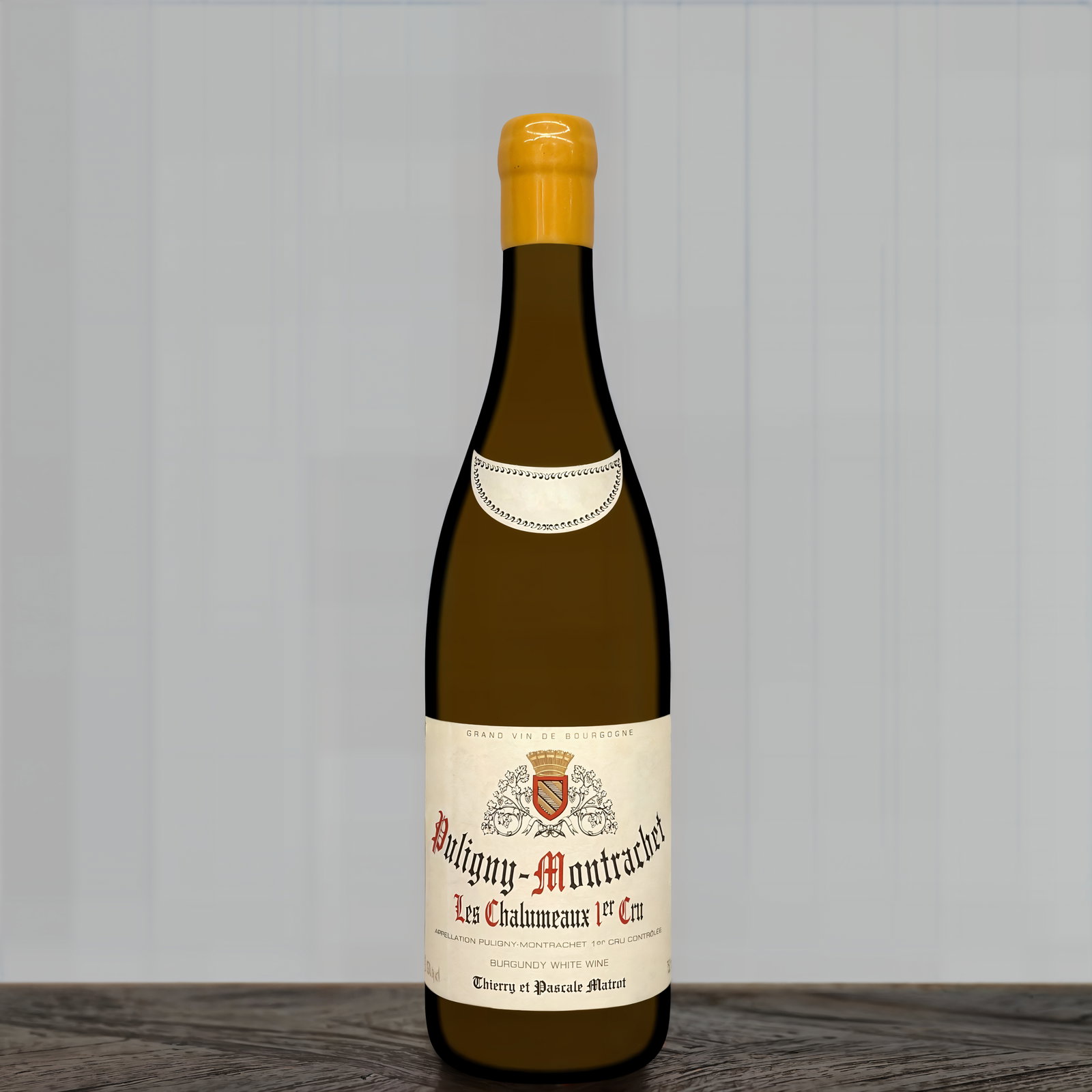 A bottle of Puligny-Montrachet wine with a cream label and gold cap sits on a wooden surface against a gray-striped background.