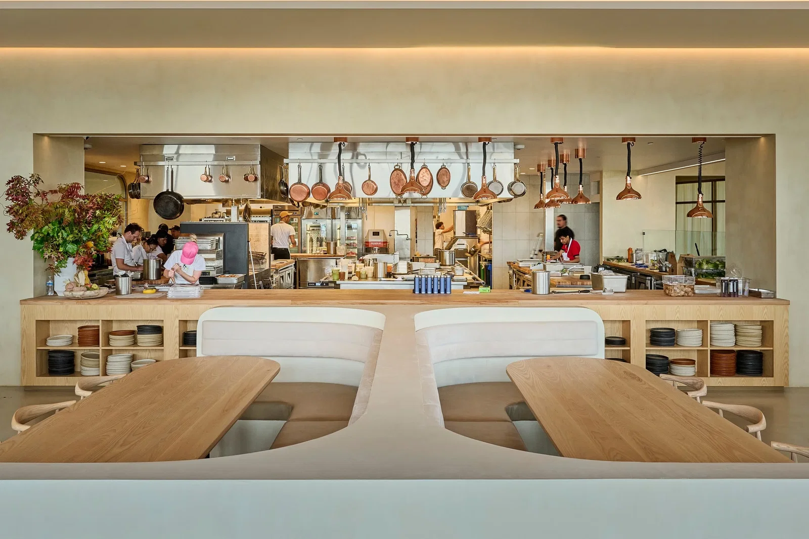 Open kitchen with chefs working under hanging copper pans. Wood tables and shelves with dishes in foreground. Bright, modern ambiance.