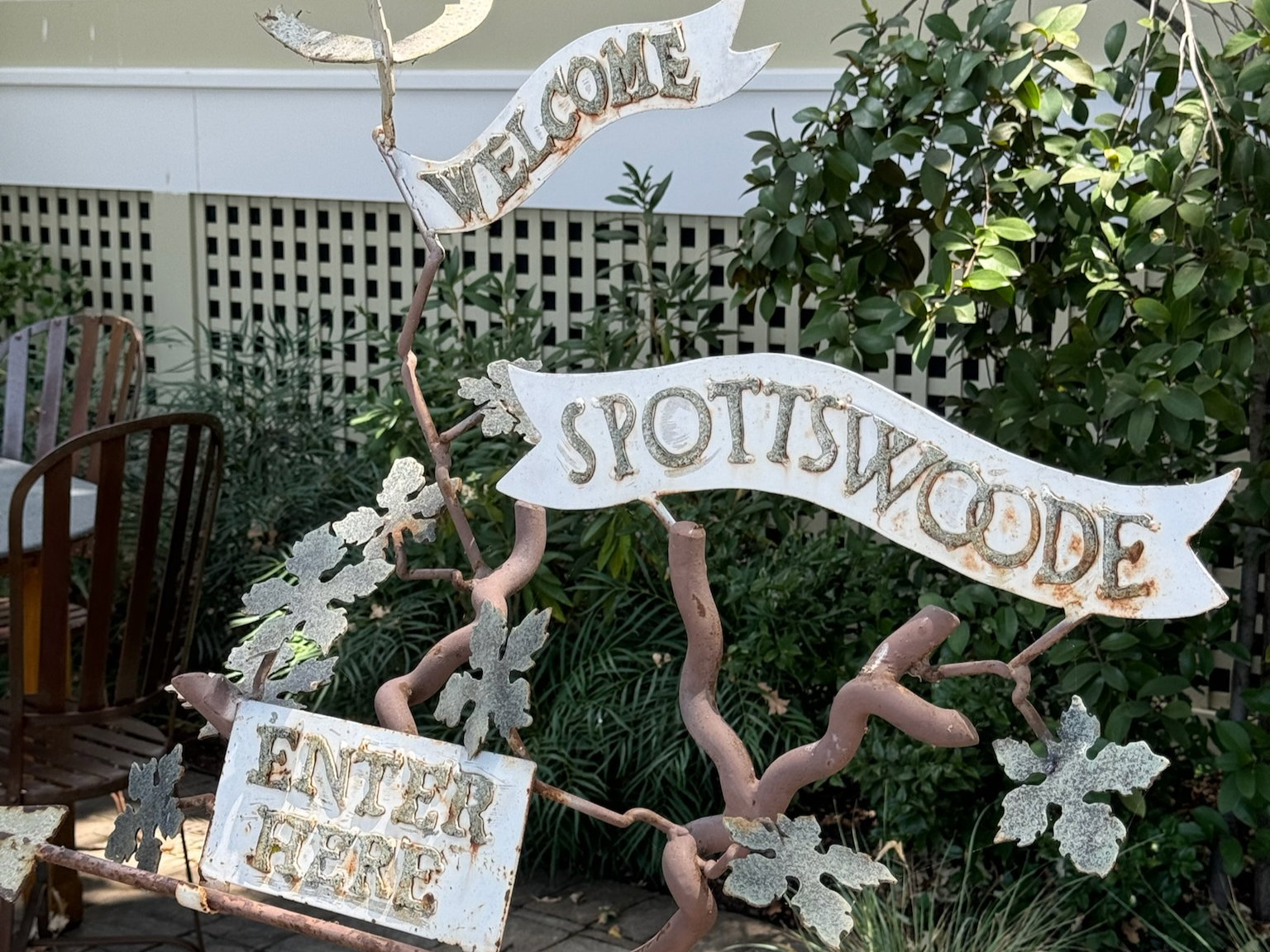 Rustic sign reading "Welcome Spottswoode Enter Here" with vines and leaves, set against a garden and lattice fence, creating a welcoming vibe.