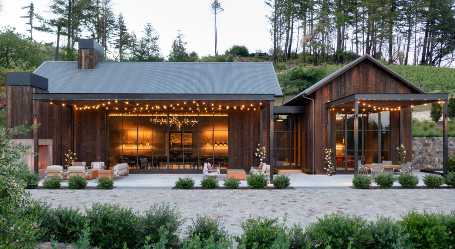 Modern wooden villa with large windows, surrounded by trees. Warm interior lighting and string lights create a cozy evening ambiance.