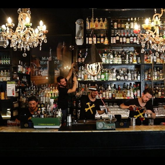 Bartenders in black shirts with yellow designs mix drinks in a dimly lit bar, adorned with chandeliers and shelves full of colorful bottles.