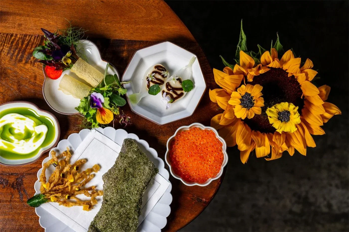 Elegant dishes with vibrant garnishes on a wooden table; bright sunflowers, salmon roe in a white bowl, and green sauce adding color.
