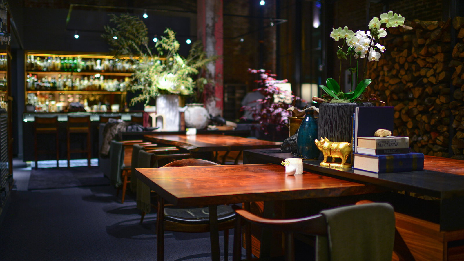 Cozy restaurant with wooden tables, plush chairs, and ambient lighting. A bar is in the background; books and plants adorn the tables.