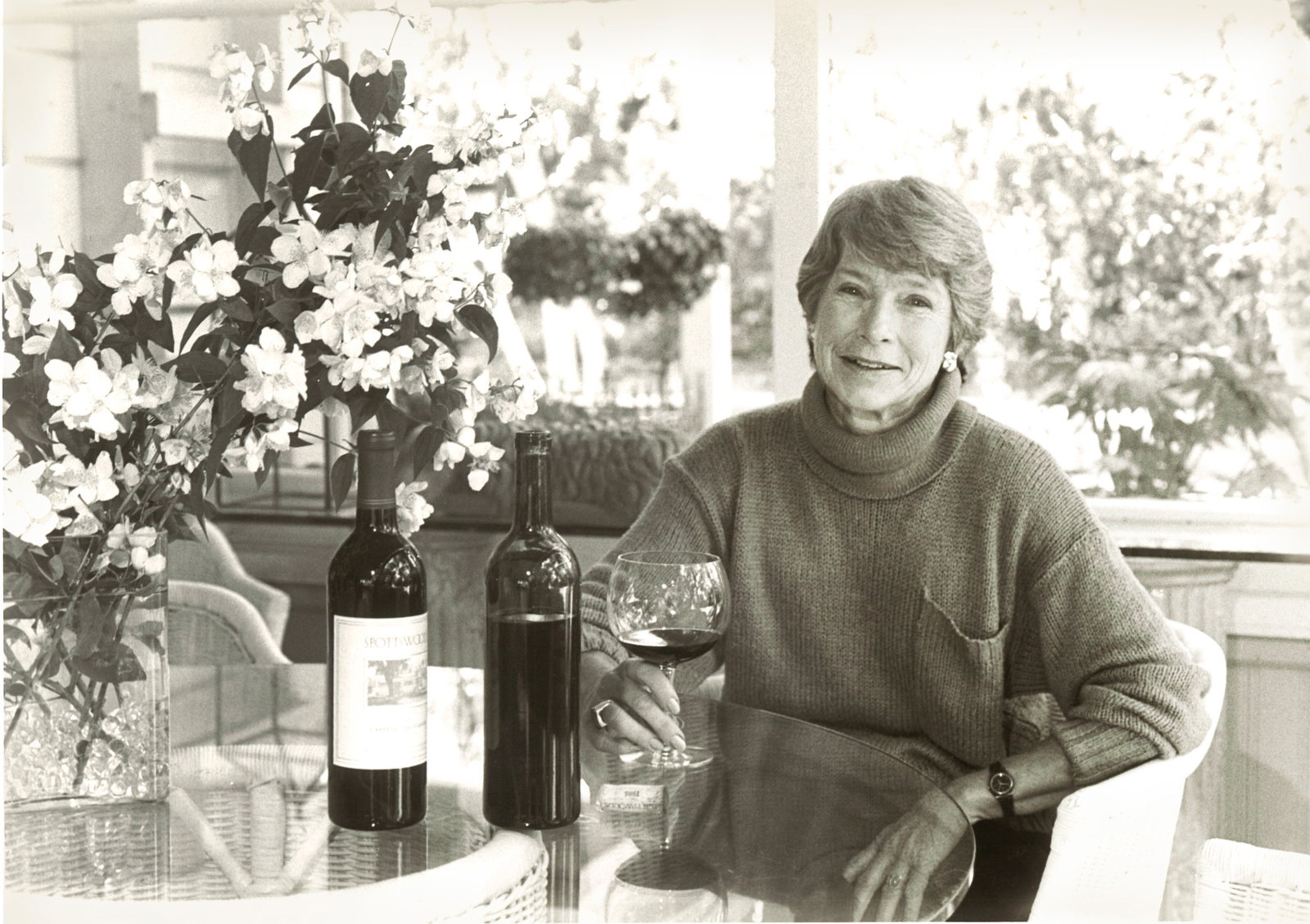 Woman smiling, sitting at a glass table with two wine bottles, holding a glass of red wine. Vase of white flowers nearby, sunny patio.